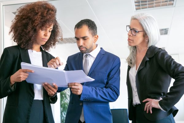 Confident office employers checking documents together. Three focused professional workers holding papers and signing statistic reports at meeting room. Teamwork, business and management concept
